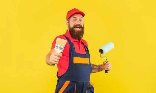 cheerful bearded man housepainter in work clothes hold paint roller and brush and on yellow background.