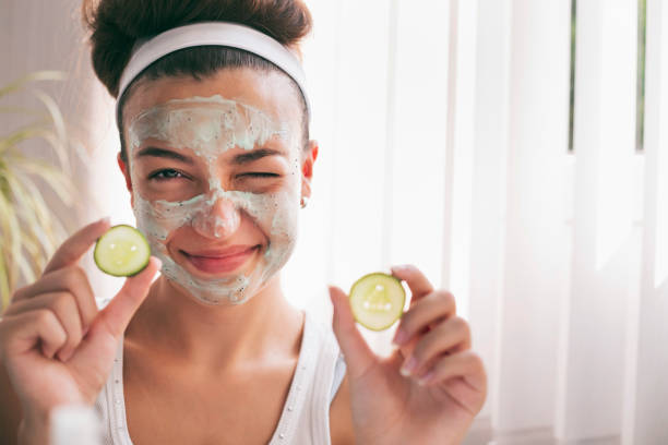 Beautiful teenage girl getting a face mask with cucumbers in home stock photo