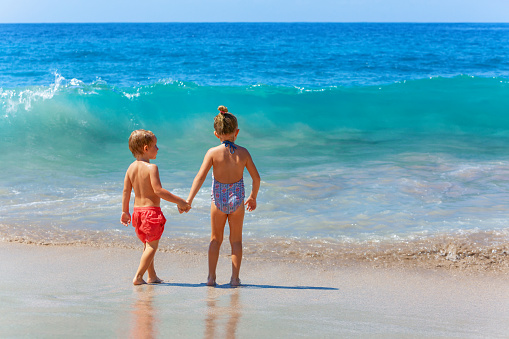 Happy kids have fun in sea surf on white sand beach. Couple of children stand in water pool with hands up. Travel lifestyle, swimming activities in family summer camp. Vacations on tropical island.