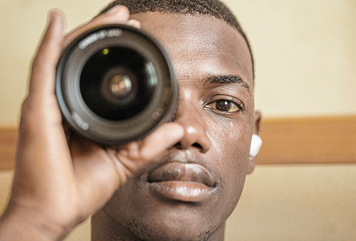 Close up of African American man holding a camera lens to his eye while looking at the camera