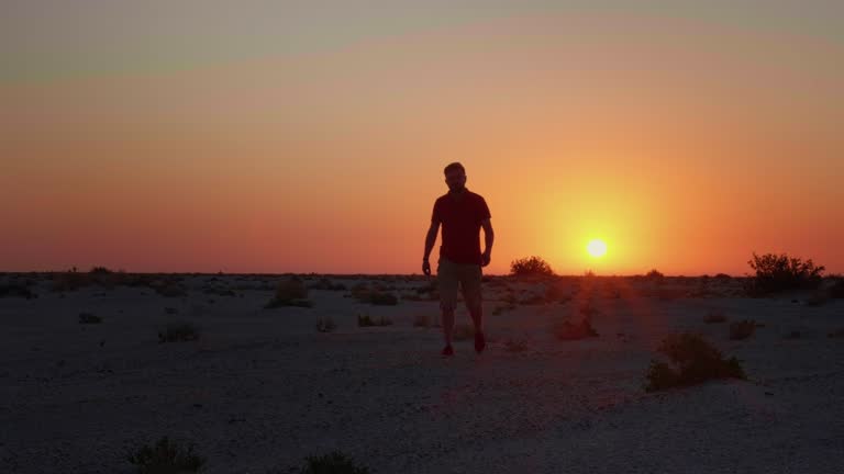 Silhouette of a man walking toward camera in desert with sunset behind.