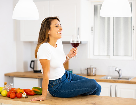 Portrait of happy smiling young Hispanic woman sitting on table in home kitchen with glass of wine