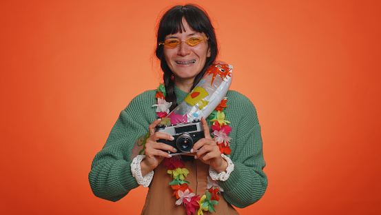 Happy young woman tourist photographer with pigtails in sunglasses taking photos on retro camera and smiling. Travel, summer holiday vacation. Latin girl traveler isolated on studio orange background