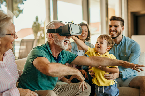 Grandfather and grandson having a great time together while using virtual reality goggles sitting on the sofa at home with the rest of the family members. Cheerful aged man wearing VR glasses with his family.