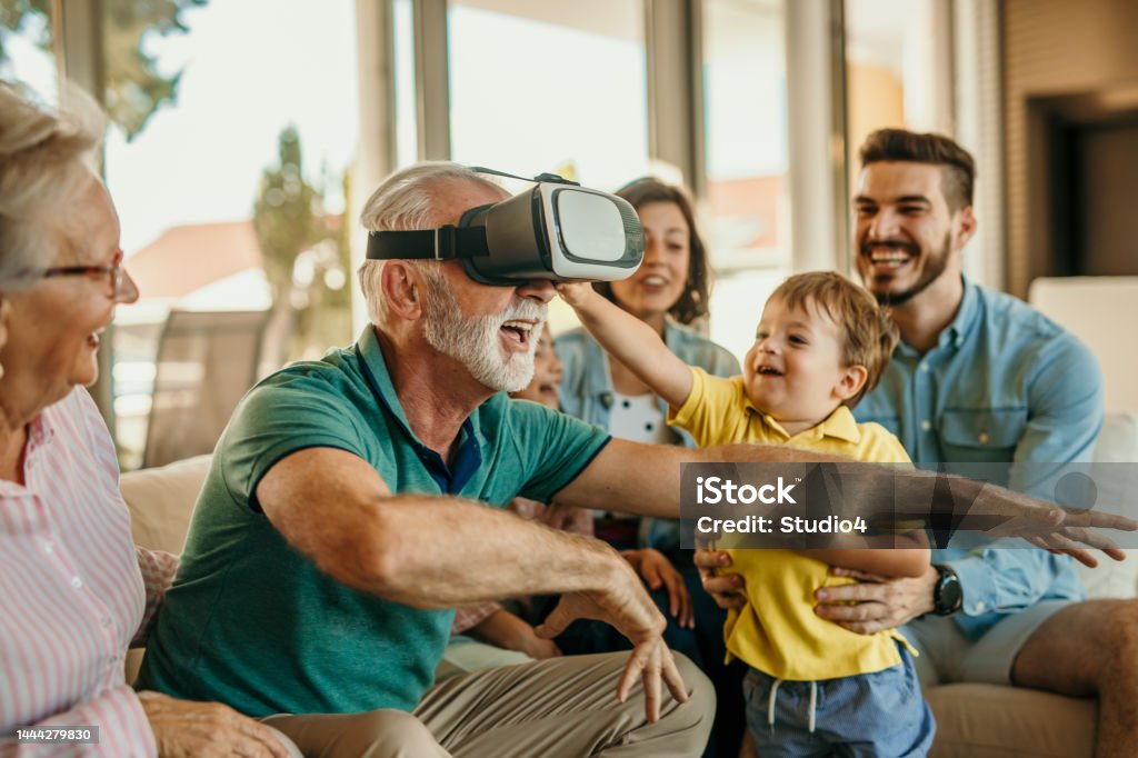 Family and VR tech Grandfather and grandson having a great time together while using virtual reality goggles sitting on the sofa at home with the rest of the family members. Cheerful aged man wearing VR glasses with his family. Virtual Reality Simulator Stock Photo