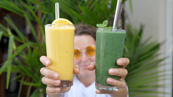 Variants of cold refreshing natural plant-based drinks. Blonde girl dancing with green and yellow cocktails with metal straws. She holds them in her hands offering to choose the one she likes best.