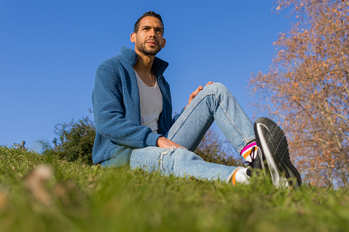 full length image of a proud gay man dressed in tight clothes sitting on the lawn and looking something away of the scene