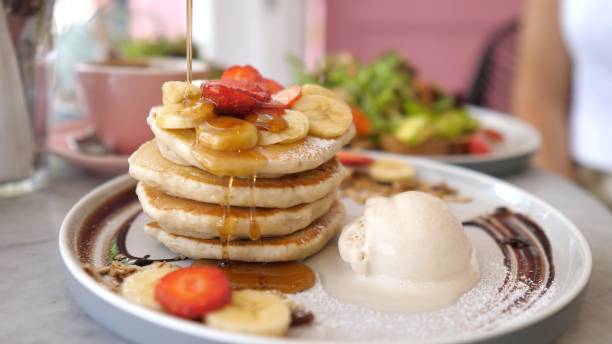 istock Sweet American breakfast. Drizzle maple syrup from a gravy boat over a stack of fluffy pancakes with a scoop of ice cream and berries. The plate is decorated with chocolate topping. 1444305091