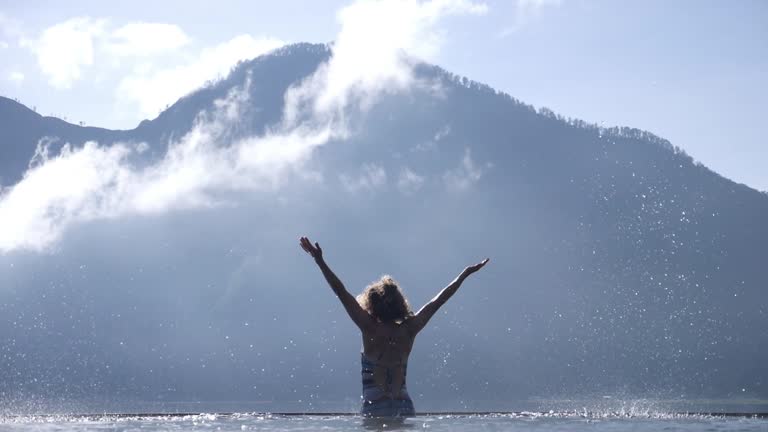 Rear view of the silhouette of a young slender woman jumping up from natural hot springs and raising her arms high. Water falls down in large drops. Mount Batur ahead.