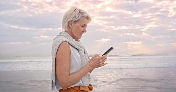 Elderly woman, phone and beach in social media, communication or chatting on a walk in the outdoors. Senior female walking on ocean coast having a online conversation or texting on mobile smartphone