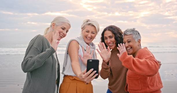 senior women, phone and video call with friends wave hello on vacation during sunset at the beach for a retirement or reunion holiday. group of  happy mature females together for online chat by sea - group of people women beach community imagens e fotografias de stock