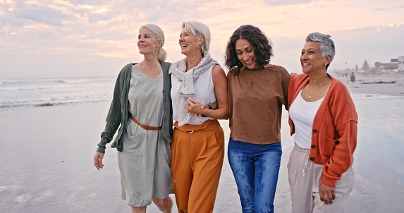 Beach, friends and vacation with a senior woman group walking on the sand by the sea or ocean. Nature, water and friendship with a mature female laughing at sunset with her retirement companions
