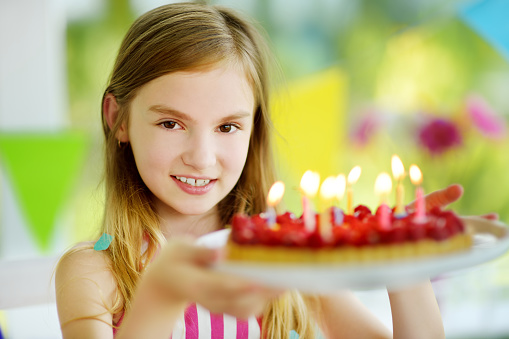 Adorable preteen girl having birthday party at home, blowing candles on birthday cake. Kids birthday party with colorful decorations, gifts and banners.