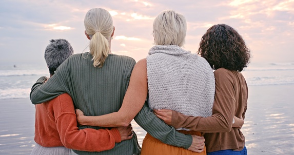 Senior women, back view and friends on beach holiday, vacation or trip outdoors. Retirement, travel and elderly group of people hug enjoying quality time together and having fun at seashore or coast.