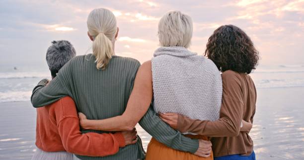 senior women, back view and friends on beach holiday, vacation or trip outdoors. retirement, travel and elderly group of people hug enjoying quality time together and having fun at seashore or coast. - group of people women beach community imagens e fotografias de stock