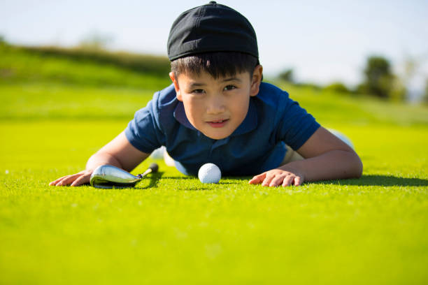 молодой восточноазиатский мальчик, лежащий на лужайке солнечного поля для гольфа в надежде на мяч для гольфа с лункой в одном - стоковое фот - golf child sport humor стоковые фото и изображения