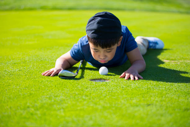 молодой восточноазиатский мальчик, лежащий на лужайке солнечного поля для гольфа, дует на мяч для гольфа в надежде, что он попадет в лунку - � - golf child sport humor стоковые фото и изображения