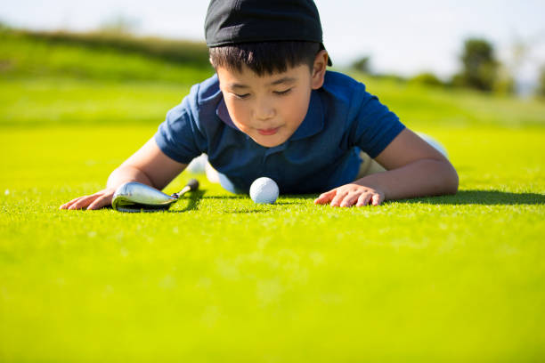 a young east asian boy lying on the lawn of a sunny golf course blowing on a golf ball hoping it will go in the hole - stock photo - golf child sport humor imagens e fotografias de stock