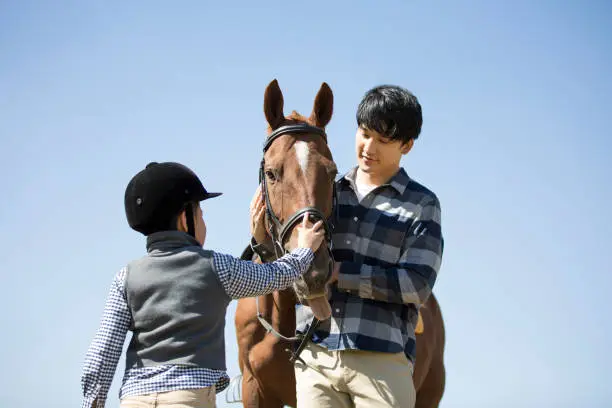 Photo of Father and son practicing horsemanship and horseback riding in horse farm under clear sky, caressing the head of horse - stock photo