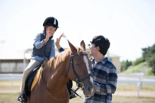 Photo of Father and son practicing horsemanship and horseback riding in horse farm, the child high-fiving his father on horseback - stock photo