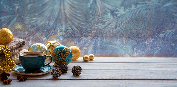 Christmas mood, holiday atmosphere. A cup of coffee, Christmas tree golden and turquoise balls, cones, star anise, cinnamon on a wooden windowsill against the background of a window covered with frosty patterns. With copy space.