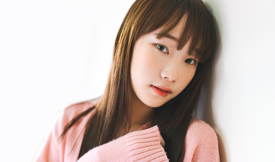 Portrait of young adult asian cute woman teen college student. Leaning on white wall attractive eye looking at camera. Long hair and wear pink sweater.