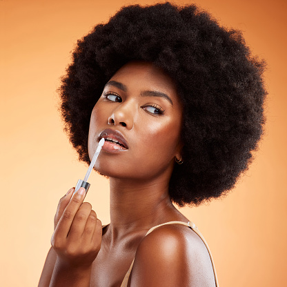 Tratamiento de brillo labial de mujer negra, afro y belleza natural para un tinte saludable, brillante y transparente. Cosméticos, aplicar y hermosa cara de modelo africana sosteniendo la herramienta de maquillaje sobre fondo naranja. photo