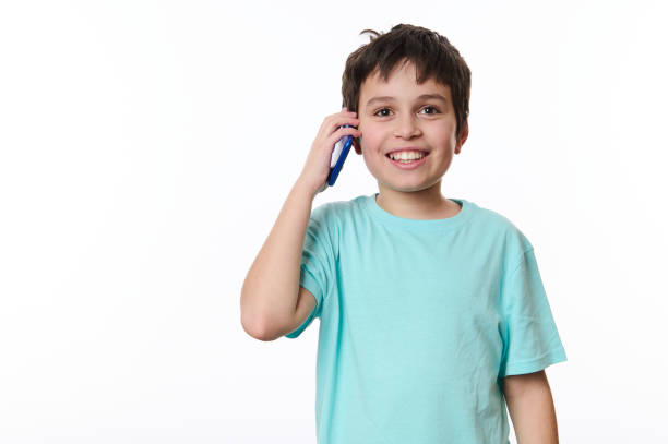Overjoyed excited dark-haired Caucasian boy, wearing turquoise t-shirt, cutely smiling a cheerful toothy smile looking at camera while talking on mobile phone, over white background with copy space