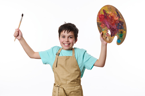 Mischievous teenage boy wearing beige apron, talented painter with paintbrush and palette of colorful paints, cutely smiling a toothy smile looking at camera, white background. Kids art development
