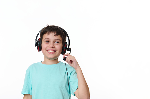 Adorable schoolboy wearing audio headset, headphones, smiling a cheerful toothy smile, looking aside at a copy space on white background. Online lesson via video link. child and technology concept
