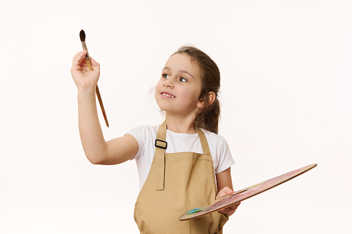 Isolated portrait on white background of a preschooler, cute baby girl holding a palette of paints, painting with a wooden brush in front of camera. Creative art development and kids entertainment