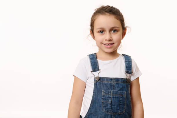 Isolated portrait on white background of a delightful baby girl, wearing blue denim overalls, smiling looking at camera stock photo