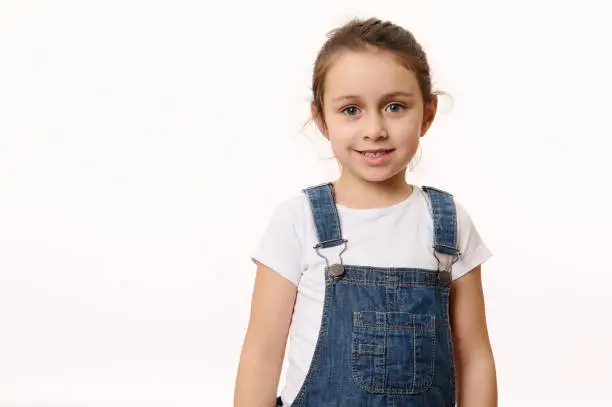 Emotional portrait isolated on white background of a delightful Caucasian baby girl, wearing blue denim overalls, happily smiling looking at camera. Beautiful kids. Children. Happy childhood concept