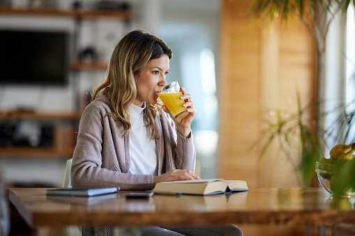 Young woman drinking orange juice and day dreaming while reading a book at home.