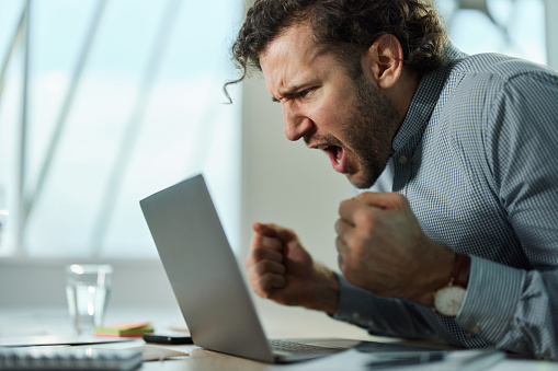 Young male entrepreneur screaming while celebrating good news received over a computer in the office.