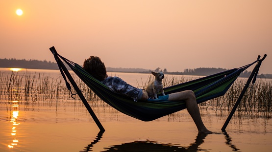 A man watches the sunset while resting in a hammock in the water with his Chihuahua.