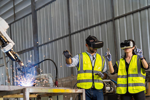 Male engineer and female working control robot arm system welding with VR glasses of virtual reality at production plant factory. Male and female worker control robot arm system welding by VR glasses
