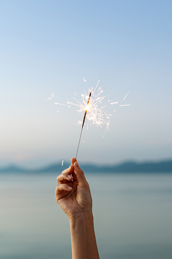 A woman's hand holds burning sparklers against the backdrop of the sea and mountains close-up bright sparks