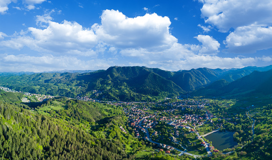 Small Bulgarian town Smolyan with houses and lake is covered with vegetation and sunbeams under clouds. Rhodope Mountains. Panorama, top view