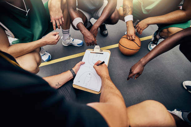 Basketball, sport and team with a sports coach talking to his team while planning tactics on a clipboard from above. Teamwork, fitness and exercise with a player and teammates listening at training baloncesto, estrategia y equipo con un entrenador deportivo hablando con un equipo mientras planifica tácticas en un portapapeles y manos. trabajo en equipo, fitness y ejercicio con un jugador y compañeros de equipo escuchando en el entrenamiento