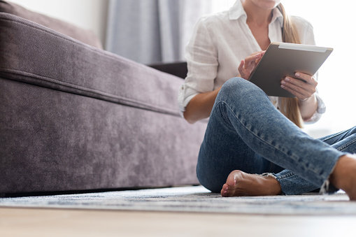Young woman in white blouse and jeans sitting on a floor at home and using a tablet. Using technology.