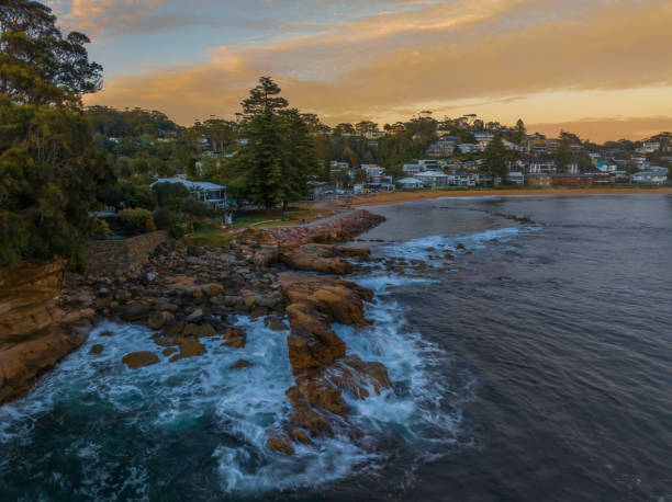 Sunrise over the ocean with rocks and coastal suburb Aerial sunrise at the seaside with rocks and gentle surf at Avoca Beach on the Central Coast, NSW, Australia. avoca beach photos stock pictures, royalty-free photos & images