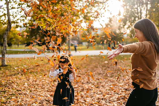 Young mother and daughter playing with a heap of autumn leaves in the park in autumn.