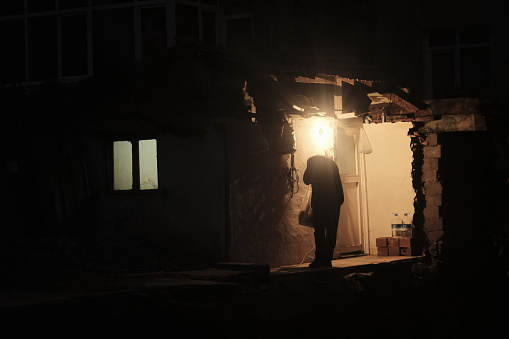 A man with a bag in front of a destroyed house. He has a lamp on his head. night and dim lights.