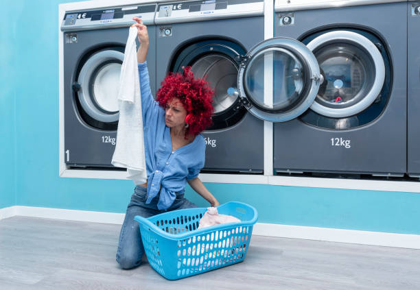 una joven latina con cabello afro rojo se sienta sacando ropa sucia de la canasta en una lavandería automática azul escuchando música con auriculares rojos. - all laundry detergent audio fotografías e imágenes de stock