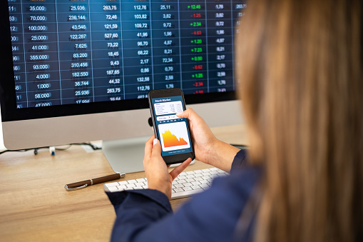 Business woman at the desk with mobile phone in the hand. Dropping stock chart on the screen.  PC monitor in the background. Stock market, office, trading and business concept.