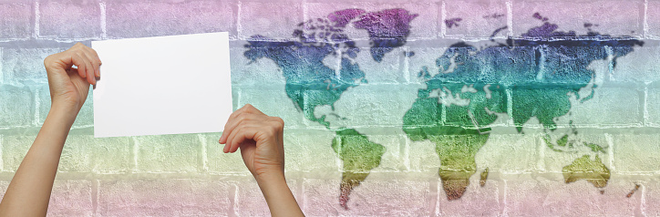 hands holding blank sign against brick wall with map of world in graduated rainbow colours and space for copy