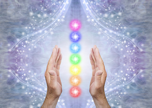 Male parallel hands with 7 rainbow coloured energy centres between against a blue flowing sparkling background and copy space