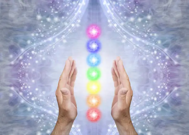 Male parallel hands with 7 rainbow coloured energy centres between against a blue flowing sparkling background and copy space