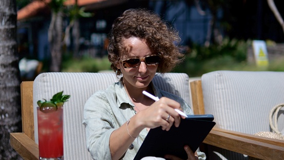 Freelance work from anywhere in the world via the Internet. Curly girl in sunglasses works as an illustrator sits on the beach in tropics with a soft drink is engaged in work using a graphics tablet